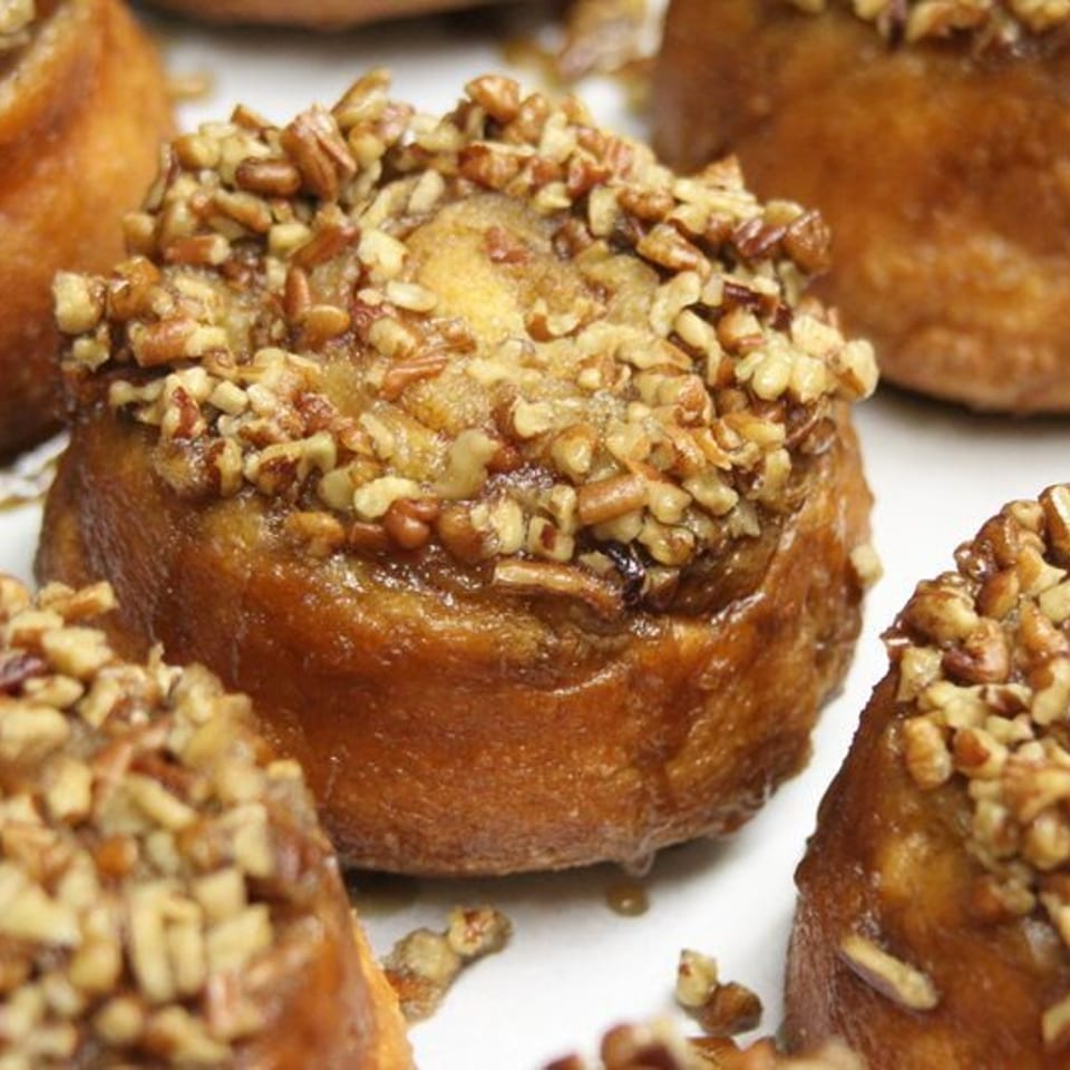 Close-up of golden pastries topped with chopped pecans, arranged closely together on a white serving surface.