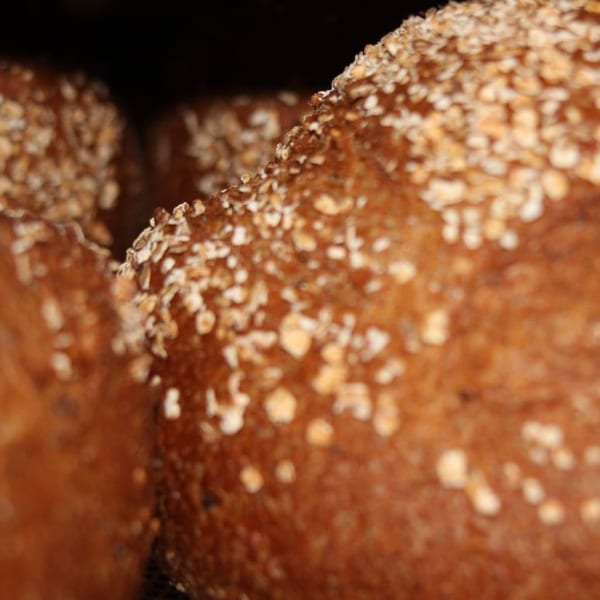 Close-up of two loaves of dark bread, sprinkled with oats, against a dark background.
